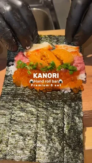 ภาพระยะใกล้ของแฮนด์โรลปลาไหลญี่ปุ่น (Unagi hand roll) ที่ห่อด้วยสาหร่ายสีเข้ม มือหนึ่งกำลังถือแฮนด์โรลนี้ ด้านบนเผยให้เห็นเนื้อปลาไหลย่างซอสสีน้ำตาลเข้มมันวาว และท็อปด้วยไข่หอยเม่น (Uni) สีส้มสดใสหลายชิ้น พร้อมผักโรยสีเขียว เมนูใหม่ล่าสุด "The Unagi" นี้เป็นส่วนหนึ่งของ Premium 5 set (1490 บาท) จากร้าน Kanori Handroll สาขา ICONSIAM โดยทางร้านระบุว่าปลาไหลมีหนังกรอบ เนื้อฉ่ำ และท็อปด้วย Uni จะเริ่มจำหน่ายตั้งแต่วันที่ 24 เมษายนนี้ มีข้อความซ้อนทับเป็นภาษาอังกฤษ "KANORI HANDROLL" และ "ICONSIAM"